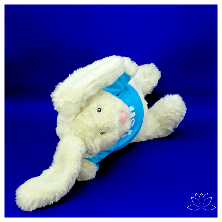 Yogahase Yon Ein weicher weißer Stoffhase mit einem blauen Shirt, liegt auf einer blauen Oberfläche.