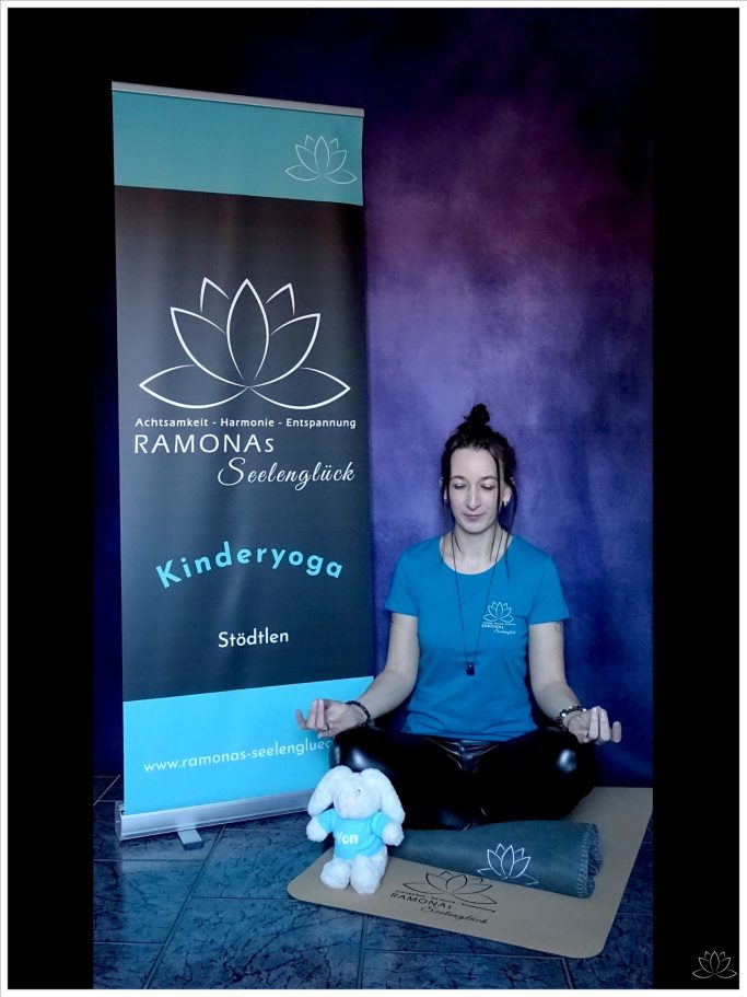 Frau im Schneidersitz mit blauem T-Shirt und schwarzer Hose meditiert mit einem Stofftier und Banner im Hintergrund.