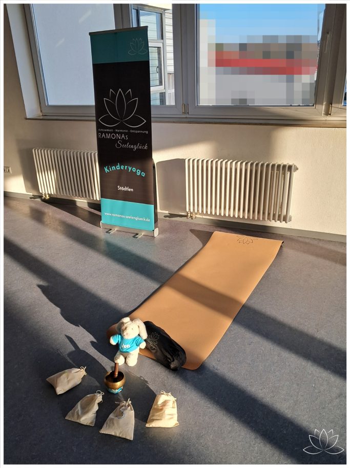 Yoga-Matte, ein Banner im Hintergrund und einige Fühlsäckchen und Klangschale auf dem Boden.