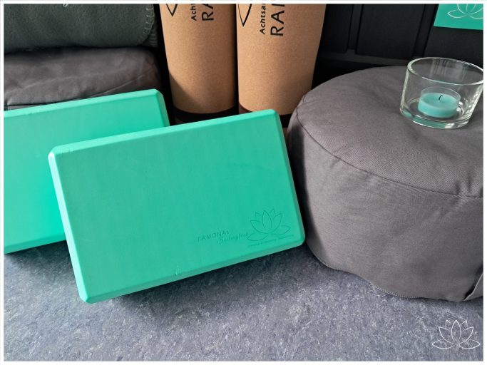 Zwei türkisfarbene Yogablöcke neben einem grauen Pouf und einem Glas auf dem Pouf.
